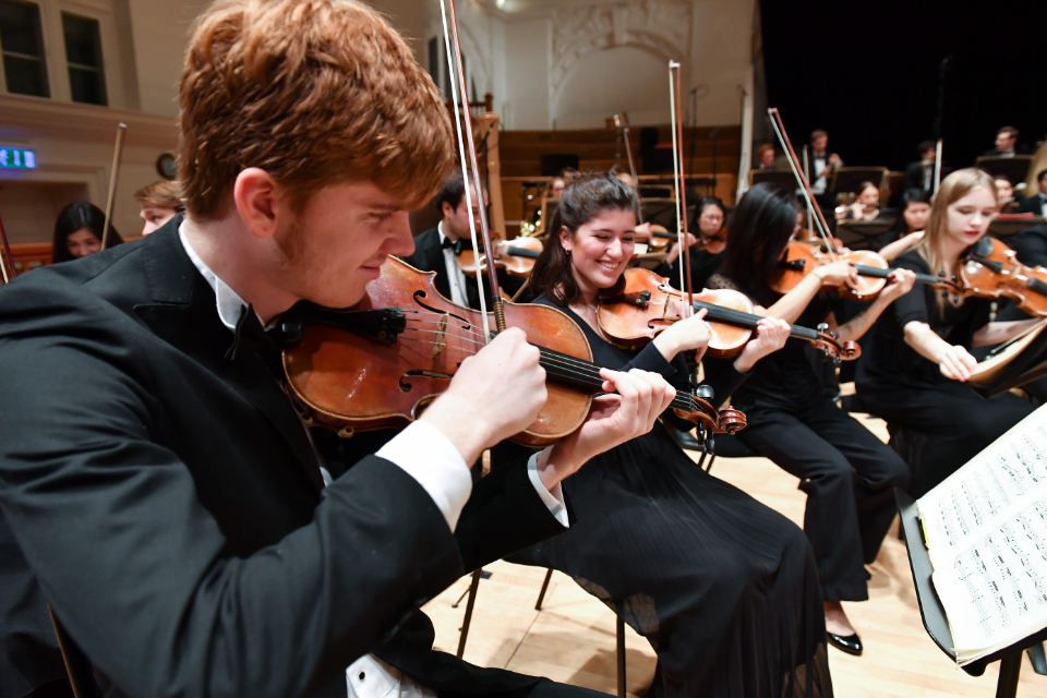 A group of students, sitting next to each other, wearing smart dress outfits, performing the violin together, in an orchestra performance.